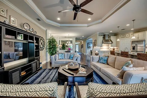 Open-plan living room with beige sofas, tray ceiling, wall TV unit, and kitchen view